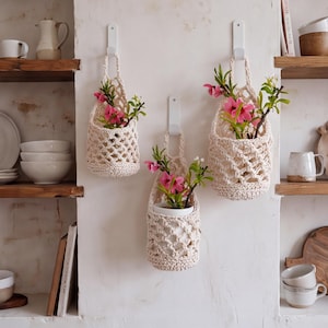 May include: Three off-white crocheted hanging baskets with a diamond lattice pattern, each suspended from a white wall hook. The baskets contain small potted plants with green leaves and bright pink flowers, displayed against a textured wall.