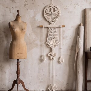 May include: A cream-colored macrame wall hanging featuring a circular top section, a horizontal wooden dowel, and a long, intricate knotted body with dangling fringe elements, displayed as a decorative piece of fiber art on a wall.