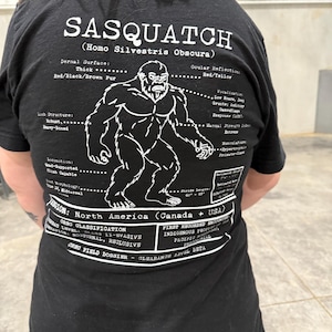 May include: A black t-shirt featuring a white graphic of a Sasquatch with scientific-style text labels. The design includes details like Dermal Surface, Limb Structure, and Locomotion, alongside the text Sasquatch (Homo Silvestris Obscura) and Orbo Field Dossier.