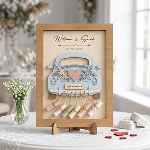 Peut inclure: Un panneau encadré en bois avec une découpe de voiture bleu clair, un cœur rose, des motifs floraux et le texte William & Sarah, 25.05.2025, et just married. Six petits rouleaux de papier sont suspendus à la voiture par de la ficelle.