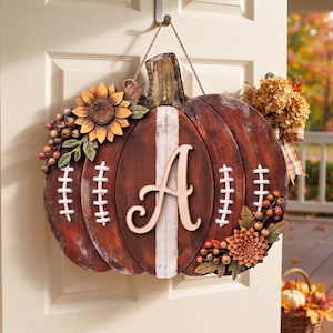 May include: A decorative door hanger shaped like a pumpkin with a brown wood-grain finish, featuring a white vertical stripe, white football-style laces, and a large letter A in the center. It is adorned with artificial sunflowers and berries.