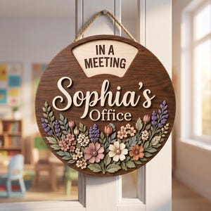 May include: A round wooden door sign with a dark wood grain finish, featuring the text "IN A MEETING" and "Sophia's Office" in raised lettering. The sign is decorated with a colourful arrangement of 3D floral elements in pink, white, and purple.
