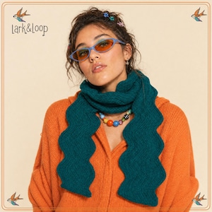 May include: A dark teal knitted scarf with a wavy, scalloped edge design. The scarf is worn around the neck, paired with an orange long-sleeved top and a necklace featuring multicolored round beads. The brand name Lark&Loop is visible.