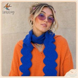May include: A bright orange knit sweater paired with a long, vibrant blue crochet scarf featuring a wavy, rippled edge. The scarf is draped around the neck, and a beaded necklace with a small heart charm is visible against the orange fabric.