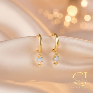 Puede incluir: Un par de pendientes de aro dorados, cada uno con una pequeña piedra opalina redonda y un dije en forma de estrella. Los pendientes se exhiben sobre una tela de satén. La joyería es de la marca "Chang Jewels".