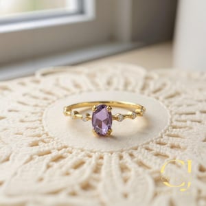 May include: A gold ring featuring an oval, purple gemstone as the centerpiece. The band is adorned with small, clear accent stones. The ring is set against a cream-colored, lace-like surface, with a window in the background.