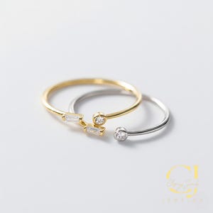 May include: Two open-ended rings. One is gold-colored with three rectangular and one round clear stone. The other is silver-colored with one round clear stone. The rings are on a white surface.