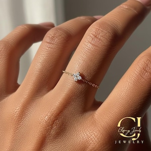 Puede incluir: Un delicado anillo de cadena dorada con un diseño de flor de cuatro pétalos con brillantes piedras preciosas blancas. El anillo se lleva en un dedo, mostrando la fina joya. El anillo es de Chany Jewels.