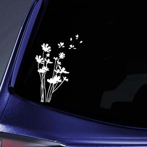 May include: White floral decal featuring a cluster of stylized flowers and several floating petals. The decal is applied to a dark surface, likely a car window, with a blue car frame visible.