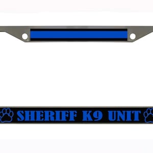 Puede incluir: Un marco de matrícula plateado con una franja negra y azul en la parte superior y las palabras "SHERIFF K9 UNIT" en azul. Huellas de patas flanquean el texto.