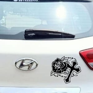 May include: Black decal of a rose, cross, and rosary beads on the rear of a white car. The decal is a stylized illustration, with the rose and cross as the central design elements. The car has a Hyundai emblem.