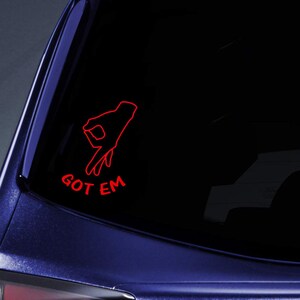 May include: A red outline of a hand making the 'gotcha' gesture, with the words 'GOT EM' below it. The sticker is on a car window, with a dark background.