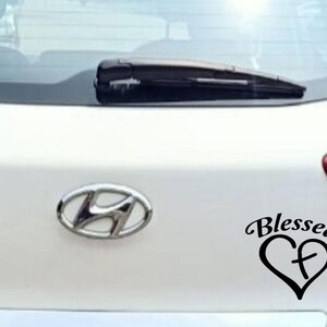 May include: A white car with a black "Blessed" heart-shaped decal. The decal features a cross inside the heart. The Hyundai logo is visible. The rear window has a wiper.