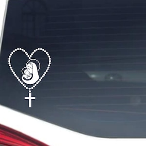 May include: A white vinyl car decal featuring a heart-shaped rosary with a depiction of the Virgin Mary and baby Jesus. A cross hangs at the bottom of the rosary. The decal is affixed to a car window.