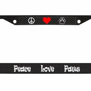 May include: Black license plate frame with a carbon fiber pattern. The top features a peace symbol, a red heart, and a paw print. The bottom has the words "Peace Love Paws" in white script.