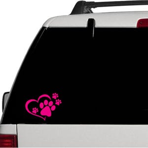 May include: A pink heart-shaped decal with paw prints on a car window. The heart contains a large paw print, with three smaller paw prints to the right. The decal is a solid pink color and is applied to the black window of a vehicle.