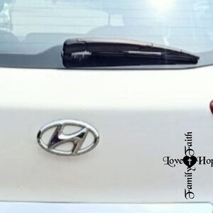 May include: A white car's rear with a Hyundai emblem and a decal featuring a cross, heart, and the words "Love, Hope, Family, Faith". The car's rear window has a wiper.