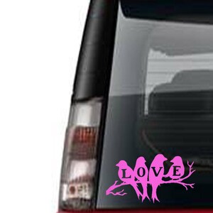 May include: Pink vinyl decal featuring the word "LOVE" with three birds perched on a branch. The decal is designed to be applied to a car window or similar surface. The image is set against the backdrop of a vehicle's rear window.
