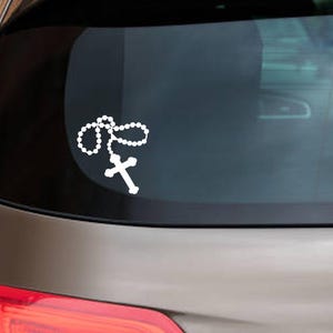 May include: White rosary bead and cross decal applied to the rear window of a car. The decal is a simple, elegant design, featuring a string of beads and a cross, suitable for religious expression.