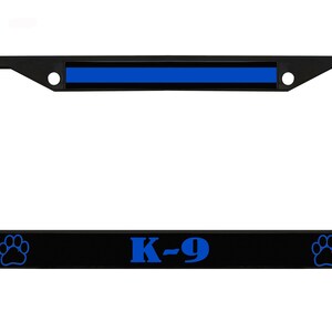 Puede incluir: Marco de matrícula negro con una franja azul en la parte superior y el texto "K-9" en azul. Cuenta con huellas de patas azules a cada lado. El marco está diseñado para una placa de matrícula estándar.