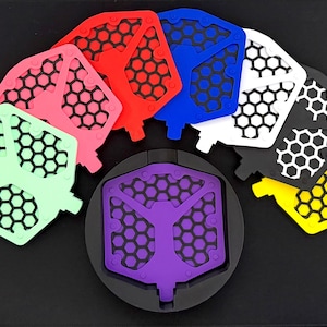 May include: A collection of colorful, geometric-patterned bicycle pedals in various colors, including pink, red, blue, white, black, yellow, and green. A purple pedal is centered on a black base. The pedals have a honeycomb design.