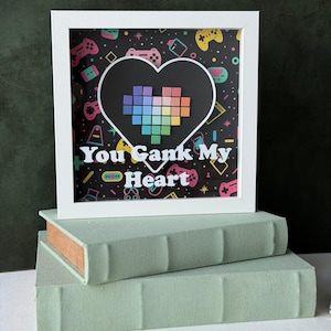 May include: A white framed art piece with the text "You Gank My Heart" over a pixelated rainbow heart. The background is black with colorful video game controller icons. The frame rests on two light green books.