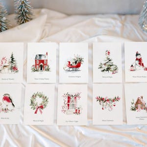 May include: A set of ten white rectangular greeting cards featuring watercolor illustrations of holiday themes including a deer, red house, sleigh, Christmas tree, snowman, robin, wreath, window, ornaments, and gingerbread house with printed text.