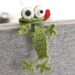 May include: A handmade, green crochet frog with large white eyes, black pupils, and a red mouth. The frog has outstretched arms and legs, appearing to hang from a gray surface. This whimsical item is a unique decorative piece.