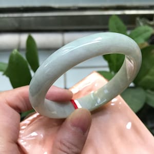 May include: A pale green jade bangle. The smooth, polished surface reflects light, showing natural colour variations. The bracelet is circular, designed for the wrist. A classic piece of jewellery, suitable for everyday wear.