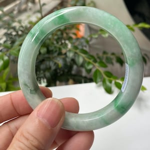 Pode incluir: Um bracelete circular e liso feito de pedra translúcida verde-clara com manchas verde-escuras espalhadas. A superfície polida tem um acabamento brilhante e apresenta uma pequena etiqueta retangular branca na lateral.