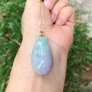 Op de afbeelding: Een jade hanger ketting met een gouden ketting. De hanger heeft een druppelvorm, met een verloop van lichtgroene en lavendelkleuren. De ketting wordt tegen een achtergrond van groen gebladerte gehouden.