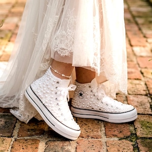 Peut inclure: Une paire de baskets montantes à plateforme blanches ornées de nombreuses petites perles blanches. Les chaussures présentent des lacets blancs, des œillets argentés et une épaisse semelle en caoutchouc blanc avec une fine rayure noire.