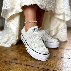 Könnte beinhalten: Weiße Converse All Star-Sneaker, verziert mit Perlen und einer durchsichtigen Schleife. Die Sneaker haben einen schwarzen Streifen auf der Sohle und eine weiße Plattform. Eine Fußkettchen aus Perlen und Strass ist ebenfalls sichtbar.