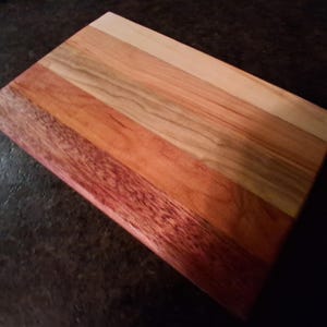 May include: A rectangular wooden cutting board with a variety of wood tones. The board is made of several strips of wood, ranging from light tan to deep red. The wood grain is visible, and the board appears to be well-crafted.