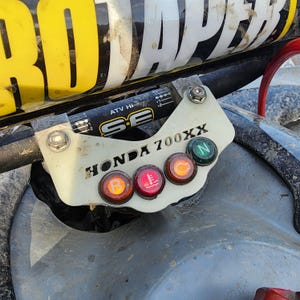 May include: Close-up of a Honda 700XX ATV control panel. The panel is off-white with the text "HONDA 700XX". It has four illuminated buttons: orange, red, orange, and green. A black bar with "ATV HI" and "SE" is visible.