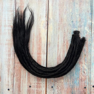 May include: A collection of black dreadlocks arranged in a curved shape. The dreadlocks are made of a dark, textured material and are displayed against a weathered, multi-toned wooden background. The ends of the dreadlocks are visible.