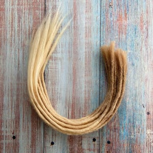 May include: A collection of blonde and light brown dreadlocks arranged in a curved shape. The dreadlocks are made of a soft, textured material and are displayed against a weathered, multi-colored wooden background. The color of the dreadlocks transitions from blonde to light brown.