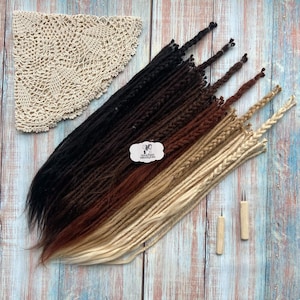 May include: An assortment of dreadlocks in a gradient of colors, from black to blonde, with two wooden crochet hooks and a cream-colored lace doily. The dreadlocks are arranged on a wooden surface with a blue and white painted design.