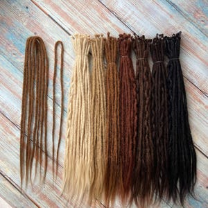 May include: An assortment of synthetic dreadlocks in various shades of brown, blonde, and black. The dreadlocks are bundled and arranged in a row, showcasing a range of colors from light to dark. The texture appears to be soft and natural-looking.
