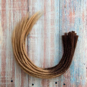 May include: A collection of brown and blonde dreadlocks arranged in a curved shape. The dreadlocks transition from a light blonde color to a dark brown at the ends. The hair is arranged on a wooden surface with a painted finish.