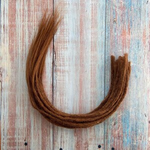 May include: A collection of brown dreadlocks arranged in a curved shape. The dreadlocks are a warm, reddish-brown color and appear to be made of a textured material. The background is a light-colored wooden surface with blue and white paint.