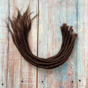 May include: A collection of brown dreadlocks arranged in a curved shape. The dreadlocks are a rich, dark brown color and appear to be made of synthetic hair. The background is a light-colored wooden surface with a mix of pink, blue, and white tones.