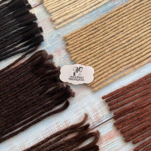 May include: An assortment of dreadlocks in various shades of brown, black, and blonde, arranged on a light blue and white wooden surface. A small, white label with the text "YOUR FIRST DREADLOCKS" is also present.