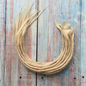 Puede incluir: Un conjunto de rastas rubias, dispuestas en forma curva. Las rastas son de color beige claro, con un aspecto ligeramente texturizado. Los extremos están deshilachados, sobre un fondo de madera con tonos azules y marrones.