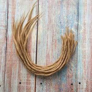May include: A collection of light brown dreadlocks arranged in a semi-circle. The dreadlocks are made of a textured material and have a natural, handmade appearance. The background is a wooden surface with a mix of blue, pink, and brown tones.