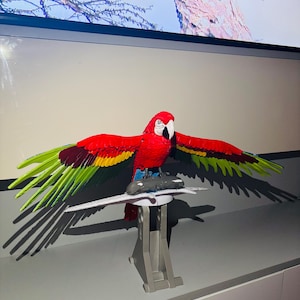 May include: A colourful parrot ornament with red, yellow, and green wings, a white face, and a black beak. The parrot is mounted on a white stand with a grey base. The figurine is displayed against a neutral background.