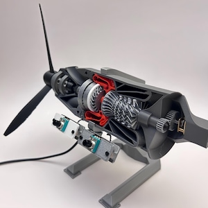 May include: A 3D-printed model of a jet engine, showing internal components. The model has a black propeller, grey housing, and red and silver internal parts. Two circuit boards are mounted on the bottom. The model is displayed on a grey stand.
