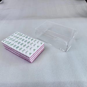 May include: A stack of white and pink tiles with green symbols and text, next to a clear acrylic box. The tiles are rectangular and stacked neatly. The box is also rectangular and transparent.