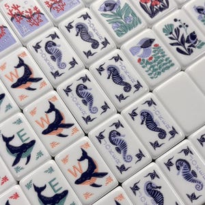 May include: White tiles with marine-themed illustrations, including seahorses, whales, and coral. Some tiles feature the word "JOKER" and numbers. The artwork is in shades of blue, orange, and red.