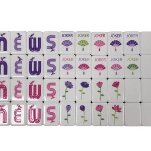 May include: A set of white Mahjong tiles. Some tiles feature the word "NEWS" in purple and pink, while others display fan and flower designs. The word "JOKER" is printed on some tiles.